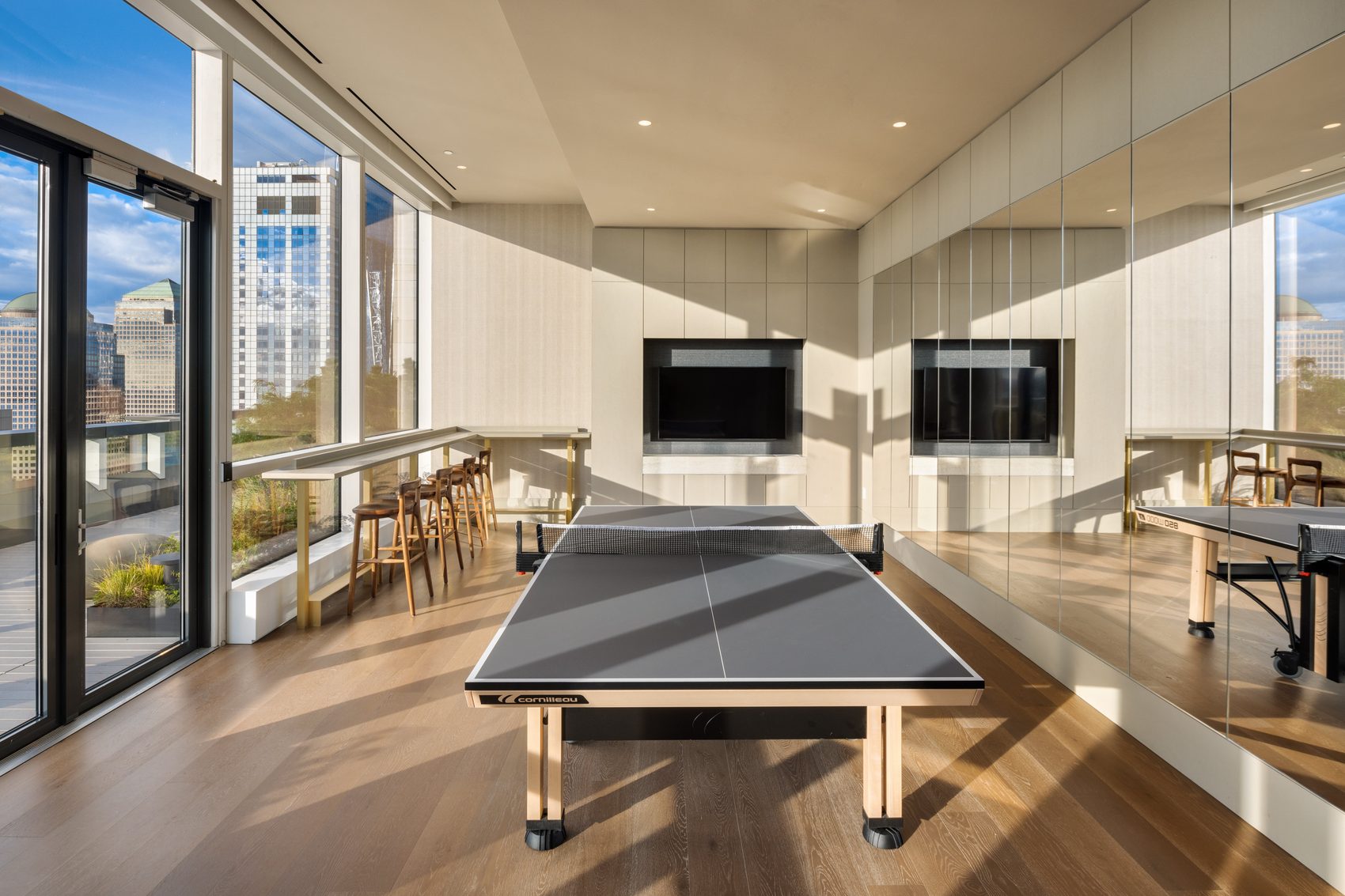 The game room with a tournament ping-pong table, mirrored wall, and floor-to-ceiling windows onto a planted terrace
